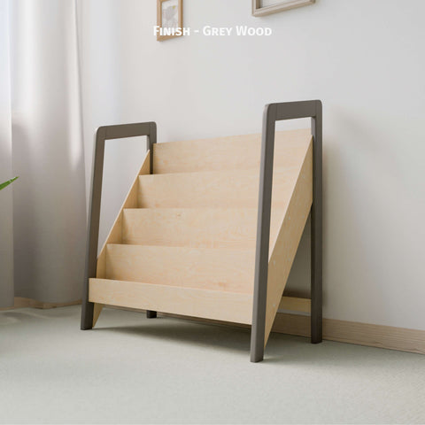 The image shows Montessori-style bookshelf made of natural wood with a grey wood finish on the frame. The bookshelf has multiple tiers designed for front-facing book display, fitting well in a light-colored, softly lit room. The minimalist design promotes a clean and organized space ideal for a child's play area or bedroom.