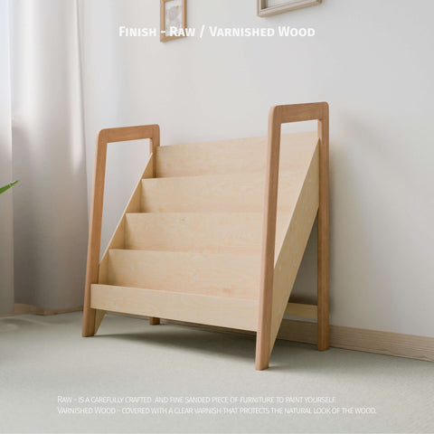 The image shows Montessori-style bookshelf made of raw or varnished natural wood. The bookshelf has multiple tiers designed for easy display and access to children's books or toys. The minimalist design fits well in a light-colored, softly lit room, promoting a clean and organized space ideal for a child's play area or bedroom.