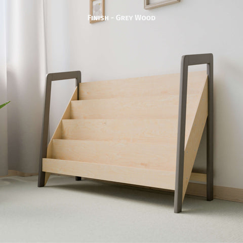 The image shows a large Montessori-style bookshelf made of natural wood with a grey wood finish on the frame. The bookshelf has multiple tiers designed for front-facing book display, fitting well in a light-colored, softly lit room. The minimalist design promotes a clean and organized space ideal for a child's play area or bedroom. The combination of natural wood and grey finish enhances the serene and inviting atmosphere, adding a contemporary touch to the room.