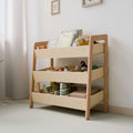  The image depicts a Montessori-style toy organizer made of natural wood, featuring three tiers of open bins. The organizer is filled with various toys and books, promoting easy access and organization for children. The design is minimalist and functional, fitting well in a light-colored, softly lit room. The open bins allow children to see and reach their toys and books easily, encouraging independence and tidy habits. 